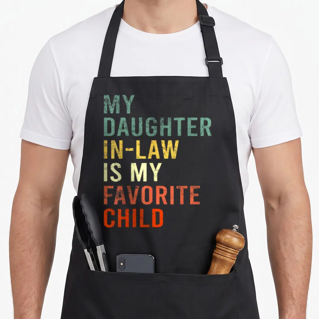 Family mom and daughter aprons - Stacked colorful text reading MY DAUGHTER IN-LAW IS MY FAVORITE CHILD in green yellow — shown on a black cotton canvas apron against a white background