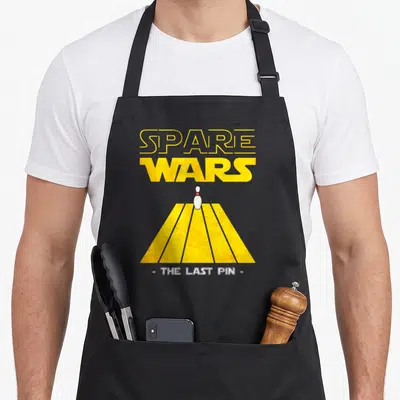 Spare pins for aprons - SPARE WARS text above white bowling pin on yellow lane with red stripes — shown on a black cotton canvas apron against a white background
