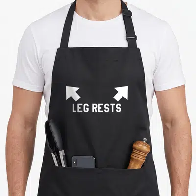 Leg dad joke apron - White LEG RESTS text with two diagonal arrows pointing up on black background — shown on a black cotton canvas apron against a white background