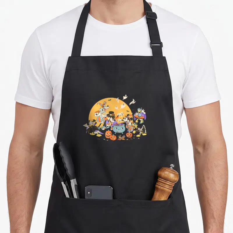 Funny friend halloween apron - Disney characters in costumes around a smiling cauldron with pumpkins and ghosts — shown on a black cotton canvas apron against a white background