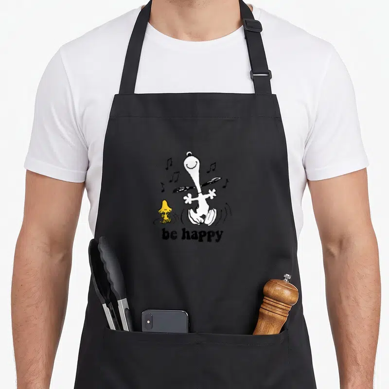 Woodstock dance apron - Cartoon white dog and yellow bird dancing with be happy text and scattered musical notes — shown on a black cotton canvas apron against a white background
