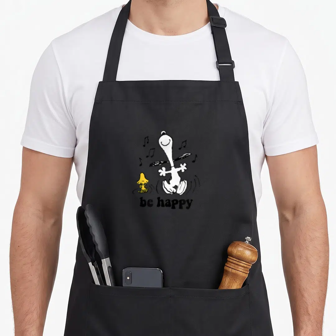 Woodstock dance apron - Cartoon white dog and yellow bird dancing with be happy text and scattered musical notes — shown on a black cotton canvas apron against a white background