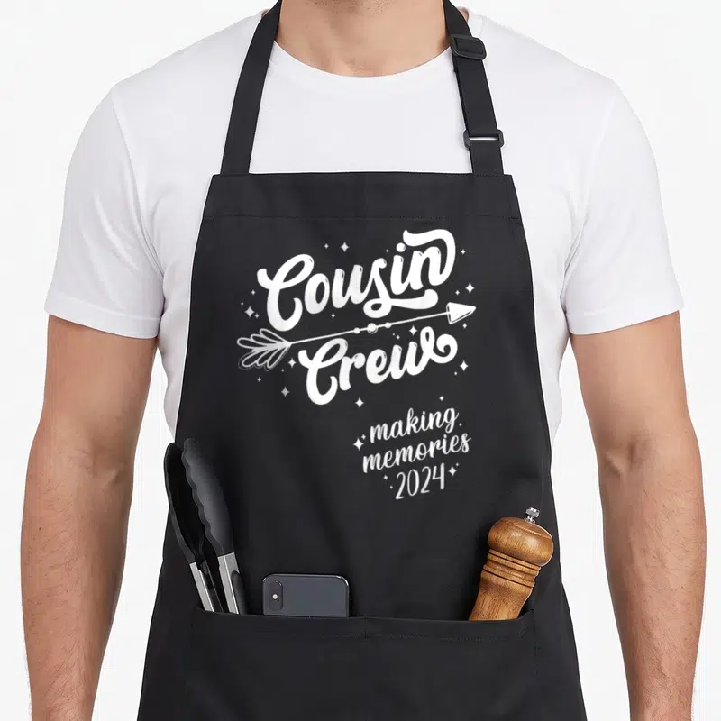 Squad memory apron - White Cousin Crew text with arrow and making memories 2024 surrounded by small stars — shown on a black cotton canvas apron against a white background