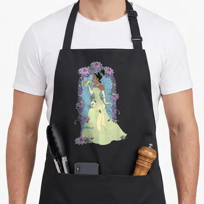 Frog floral apron - Princess in yellow gown holding green frog surrounded by purple water lilies and blue swirl patterns — shown on a black cotton canvas apron against a white background