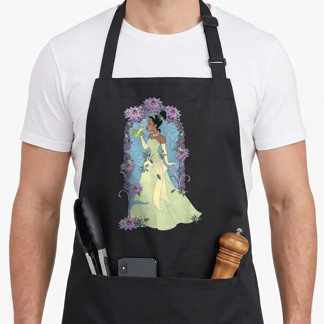 Frog floral apron - Princess in yellow gown holding green frog surrounded by purple water lilies and blue swirl patterns — shown on a black cotton canvas apron against a white background