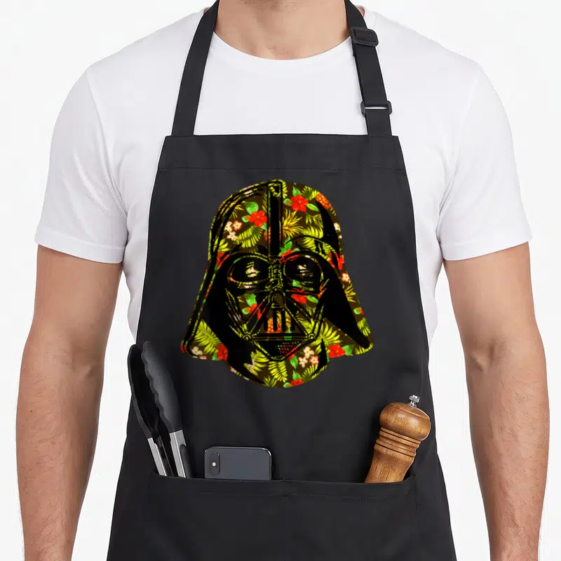 Cute helmet hawaiian apron - Darth Vader helmet silhouette filled with red and green tropical floral pattern on black — shown on a black cotton canvas apron against a white background