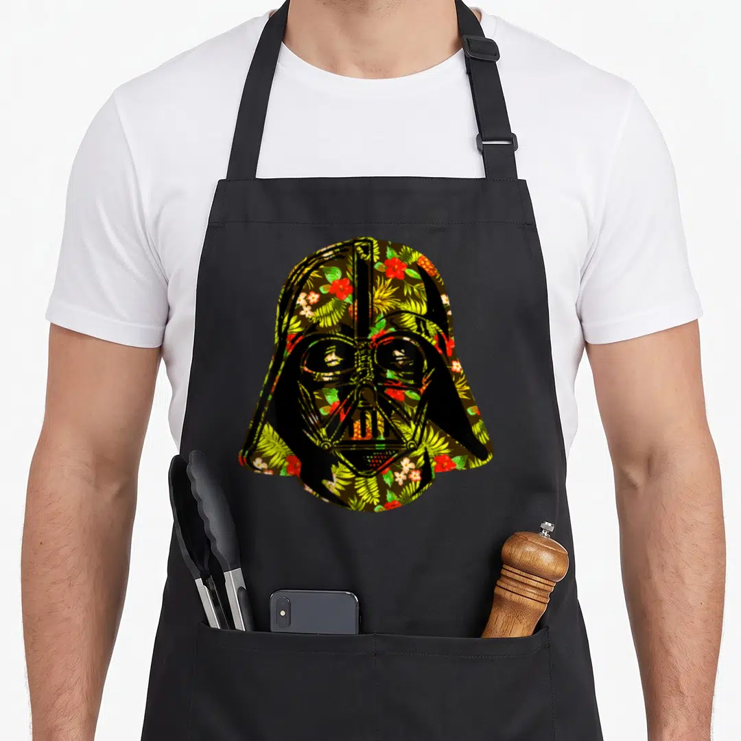 Cute helmet hawaiian apron - Darth Vader helmet silhouette filled with red and green tropical floral pattern on black — shown on a black cotton canvas apron against a white background