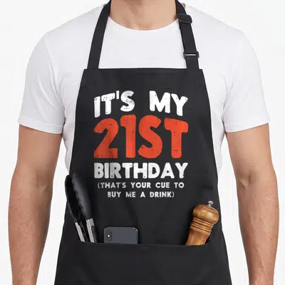 Cue birthday apron - Distressed white and red typography reading Its My 21st Birthday That's Your Cue Buy Me A Drink — shown on a black cotton canvas apron against a white background