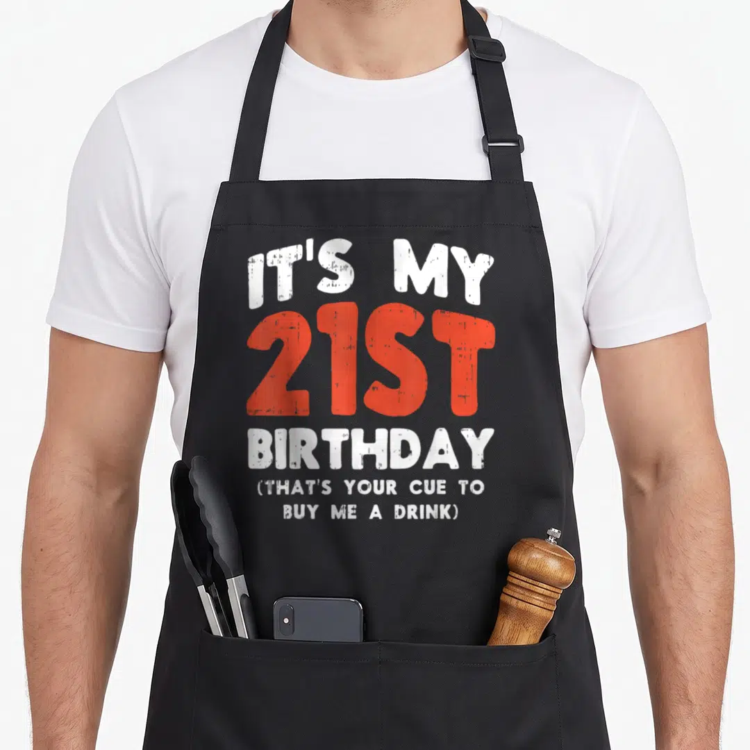 Cue birthday apron - Distressed white and red typography reading Its My 21st Birthday That's Your Cue Buy Me A Drink — shown on a black cotton canvas apron against a white background