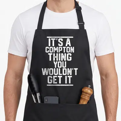 Compton family aprons - Distressed white text IT'S A COMPTON THING YOU WOULDN'T GET IT framed by two horizontal lines — shown on a black cotton canvas apron against a white background