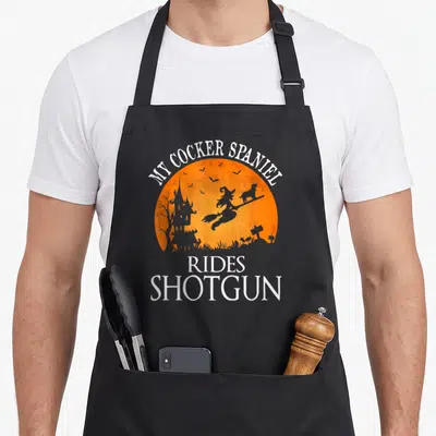 Ride cocker spaniel apron - Witch and dog silhouette on orange moon with text MY COCKER SPANIEL RIDES SHOTGUN — shown on a black cotton canvas apron against a white background