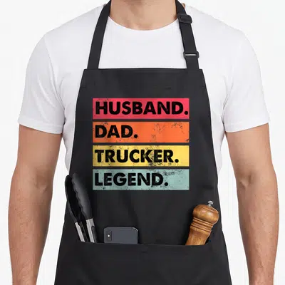 Husband truck apron - Stacked colored bars reading Husband Dad Trucker Legend in black block letters — shown on a black cotton canvas apron against a white background
