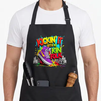 Sneaker family reunion aprons - Purple sneaker with KICKIN' IT WITH MY KIN FOLK text, boombox, and paint splatters — shown on a black cotton canvas apron against a white background