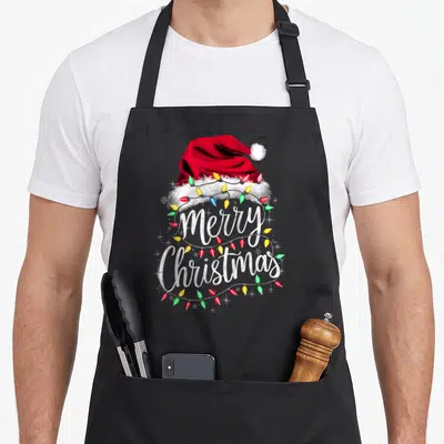 Merry santa aprons - Red Santa hat with colorful lights and Merry Christmas text in cartoon style — shown on a black cotton canvas apron against a white background
