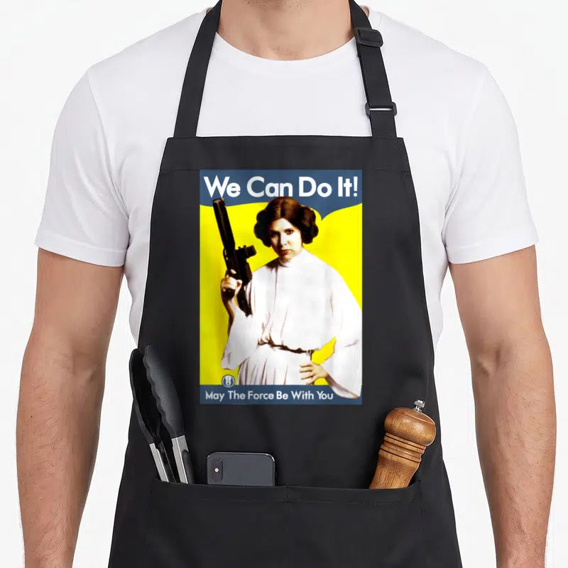 Funny poster princess apron - Retro woman in white robes holding a black blaster against yellow background with blue — shown on a black cotton canvas apron against a white background