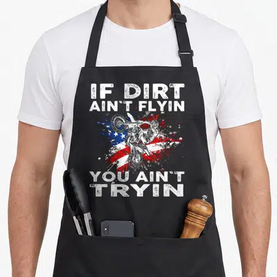 Dirtbike us aprons - Motocross rider jumping over distressed red white and blue flag with bold white text — shown on a black cotton canvas apron against a white background