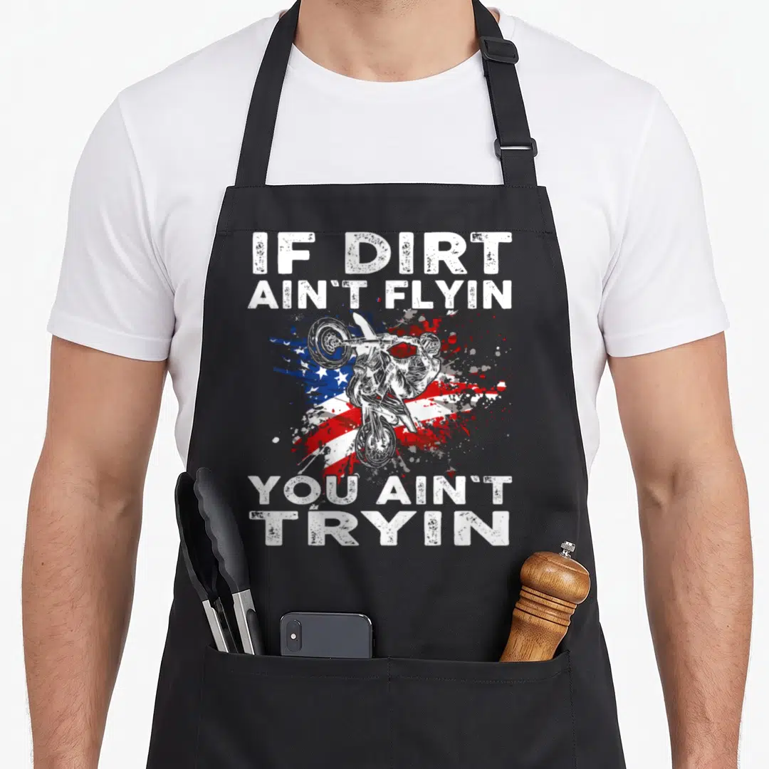 Dirtbike us aprons - Motocross rider jumping over distressed red white and blue flag with bold white text — shown on a black cotton canvas apron against a white background
