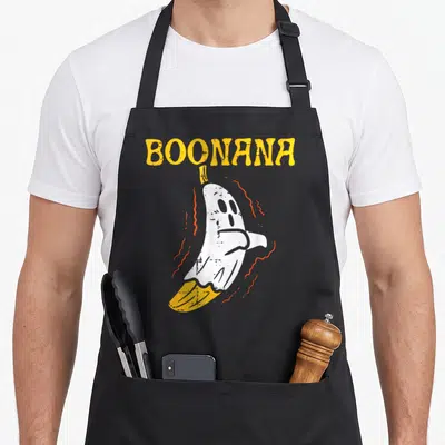 Boonana banana apron - Yellow BOONANA text above white banana ghost with orange squiggles — shown on a black cotton canvas apron against a white background