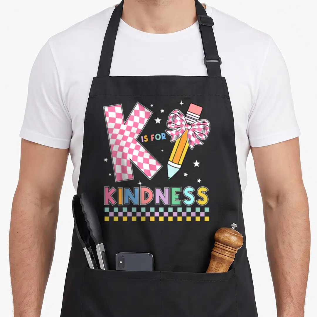 Kindness teacher apron - Pink checkered K, pencil with bow, IS FOR KINDNESS text, and checkered border — shown on a black cotton canvas apron against a white background