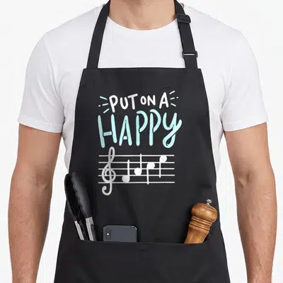 Face music teacher apron - PUT ON A HAPPY text above white musical notes on a staff in light blue — shown on a black cotton canvas apron against a white background