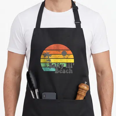 Salty summer aprons - Retro sunset with orange yellow green grey stripes, palm trees, birds, and Salty lil' Beach text — shown on a black cotton canvas apron against a white background