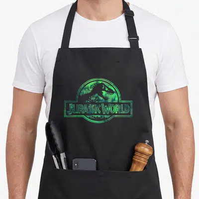 Jurassic watercolor apron - Dinosaur silhouette inside circle and rectangle with JURASSIC WORLD text in green watercolor — shown on a black cotton canvas apron against a white background