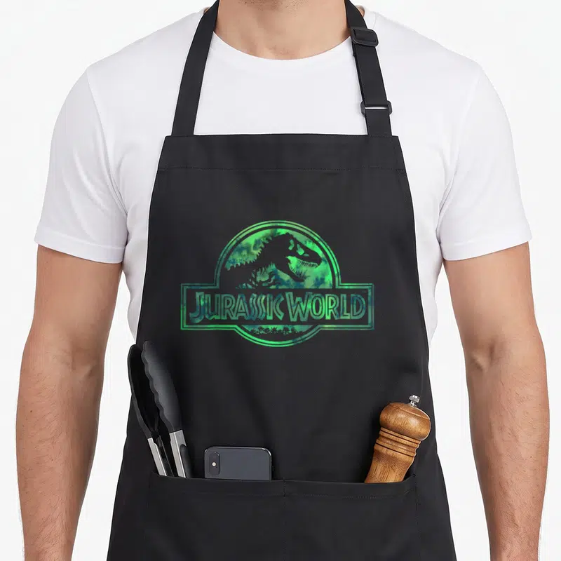 Jurassic watercolor apron - Dinosaur silhouette inside circle and rectangle with JURASSIC WORLD text in green watercolor — shown on a black cotton canvas apron against a white background