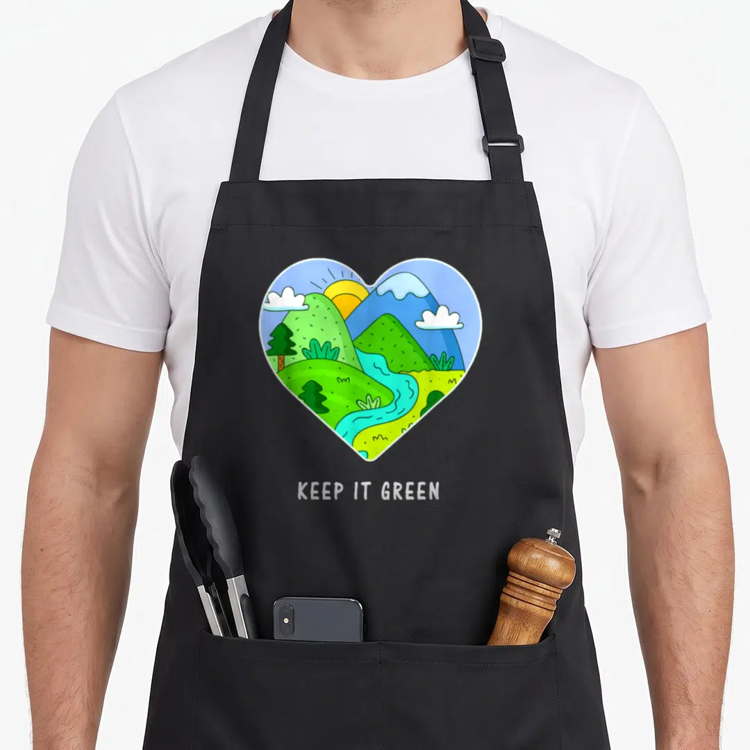Save earth day apron - Heart shaped cartoon landscape with mountains river and sun above KEEP IT GREEN text — shown on a black cotton canvas apron against a white background