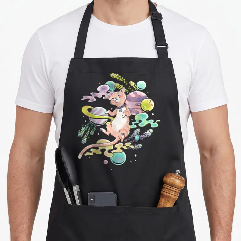 Celestial goose apron - Peach cat floating among colorful planets with COSMIC collar tag in cartoon style — shown on a black cotton canvas apron against a white background