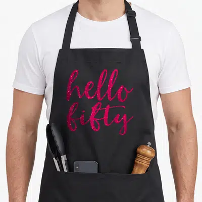 Fifty 50th birthday apron - Pink glitter script reading hello fifty stacked vertically — shown on a black cotton canvas apron against a white background