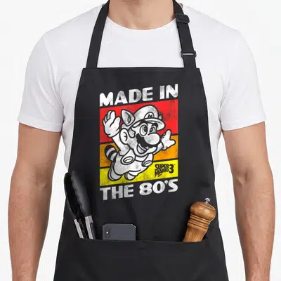 80s holiday apron - Cat Mario character over red orange and yellow stripes with retro text — shown on a black cotton canvas apron against a white background