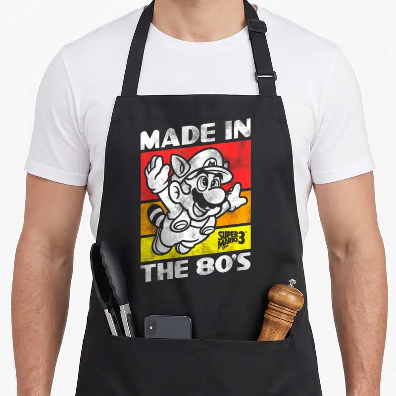 80s holiday apron - Cat Mario character over red orange and yellow stripes with retro text — shown on a black cotton canvas apron against a white background