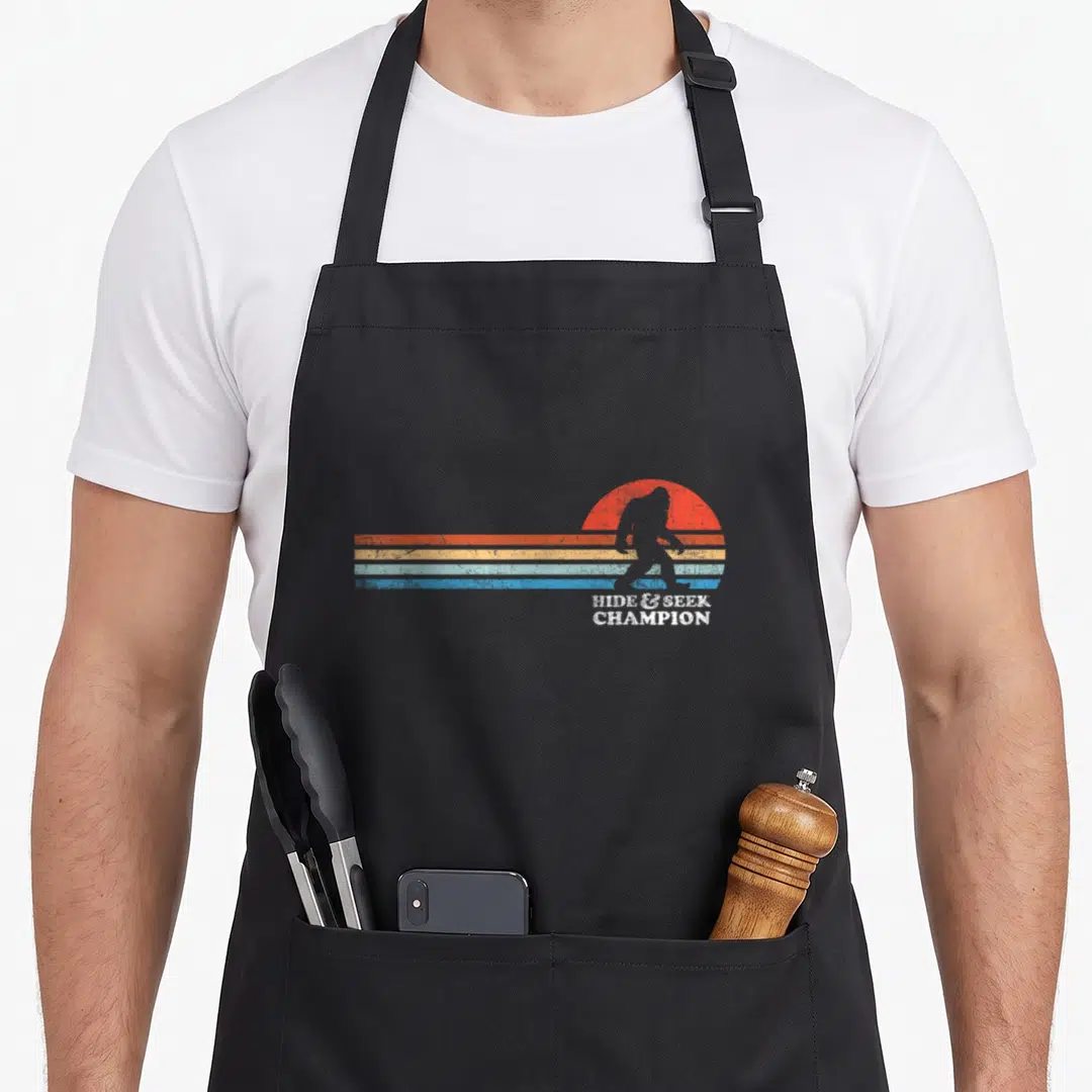 Vintage hide bigfoot apron - Retro Bigfoot silhouette over red sun and colored stripes with Hide Seek Champion text — shown on a black cotton canvas apron against a white background