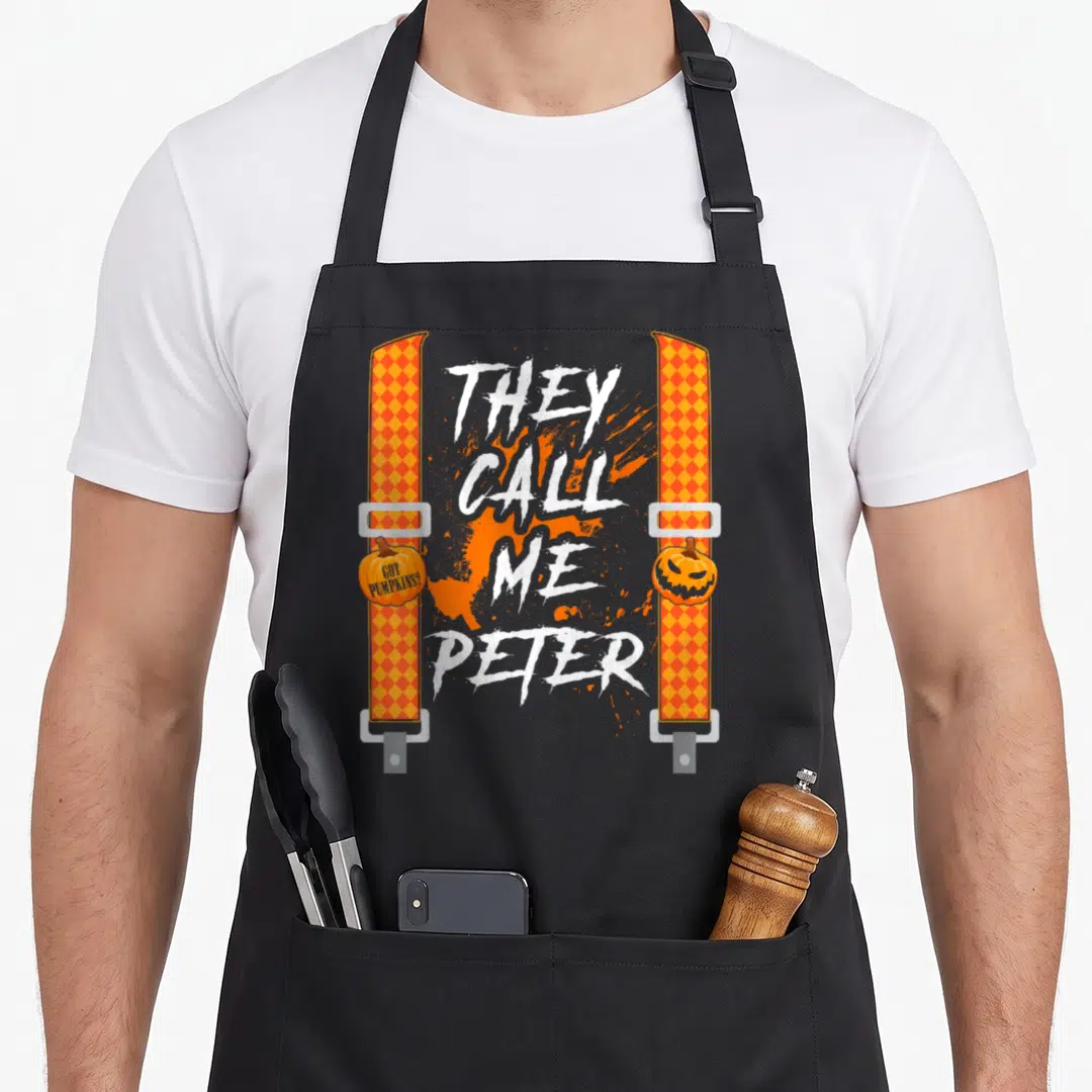 They halloween apron - Orange suspenders with They Call Me Peter text and pumpkin graphics on distressed background — shown on a black cotton canvas apron against a white background