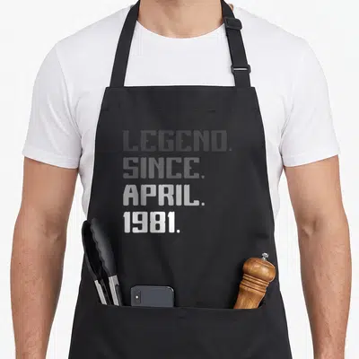 Legend april apron - Stacked white text reading LEGEND SINCE APRIL 1981 on grey background — shown on a black cotton canvas apron against a white background