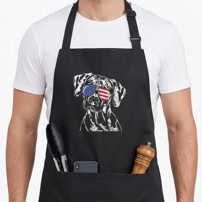 Proud rhodesia apron - Distressed white dog sketch wearing sunglasses with red white and blue flag pattern lenses — shown on a black cotton canvas apron against a white background