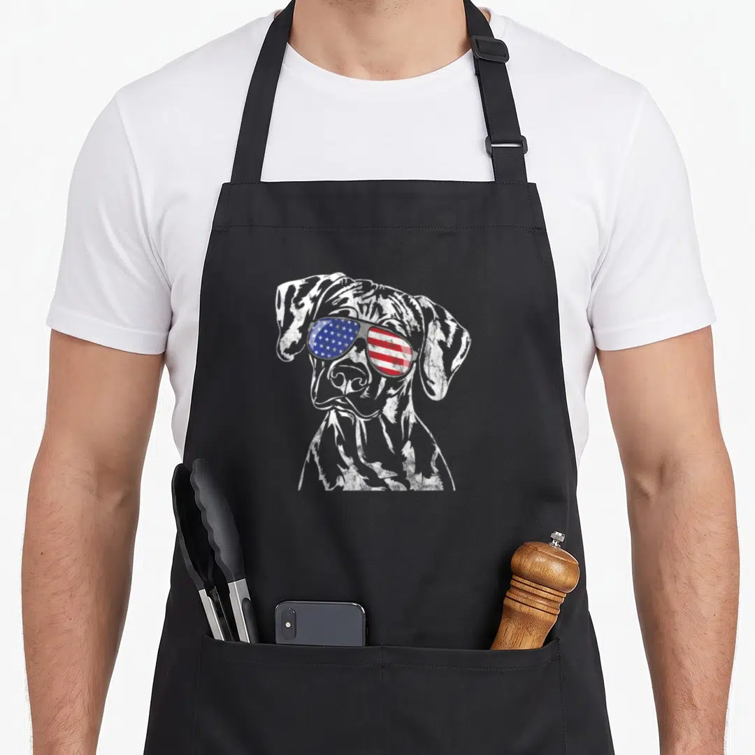 Proud rhodesia apron - Distressed white dog sketch wearing sunglasses with red white and blue flag pattern lenses — shown on a black cotton canvas apron against a white background