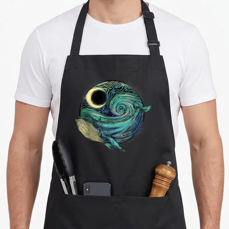 Environmentalism whale apron - Teal humpback whale swimming in front of yellow moon and swirling blue patterns inside — shown on a black cotton canvas apron against a white background