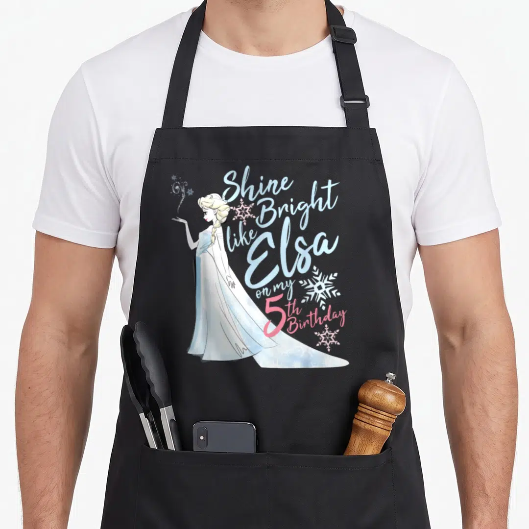 Bright birthday apron - Elsa character with Shine Bright like 5th Birthday text and snowflakes in blue and pink — shown on a black cotton canvas apron against a white background