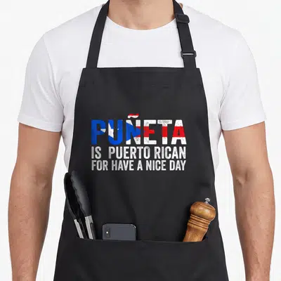 Eta beautiful apron - Stacked text Puñeta is Puerto Rican for have a nice day in flag colors — shown on a black cotton canvas apron against a white background