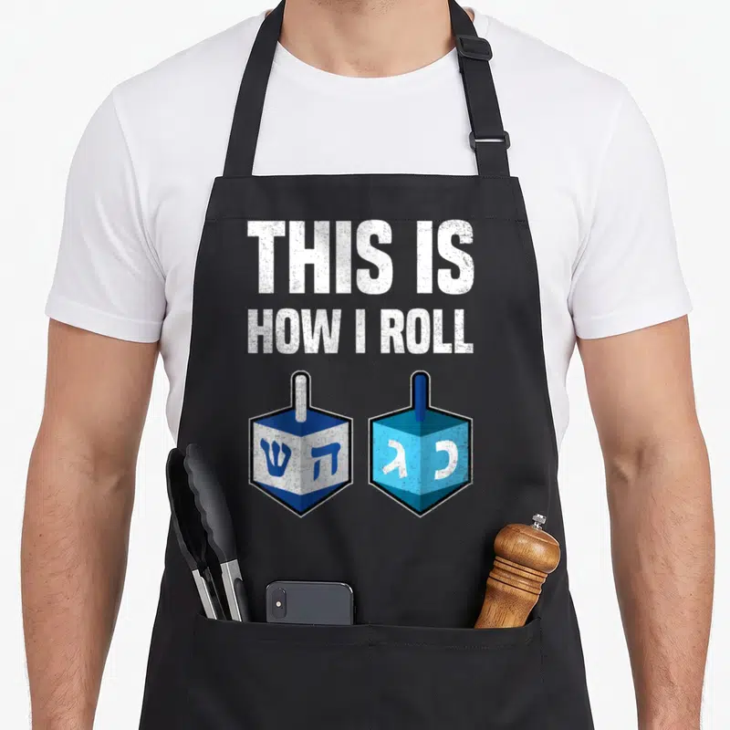Roll hanukkah apron - THIS IS HOW I ROLL text above two blue and white dreidels with Hebrew letters — shown on a black cotton canvas apron against a white background