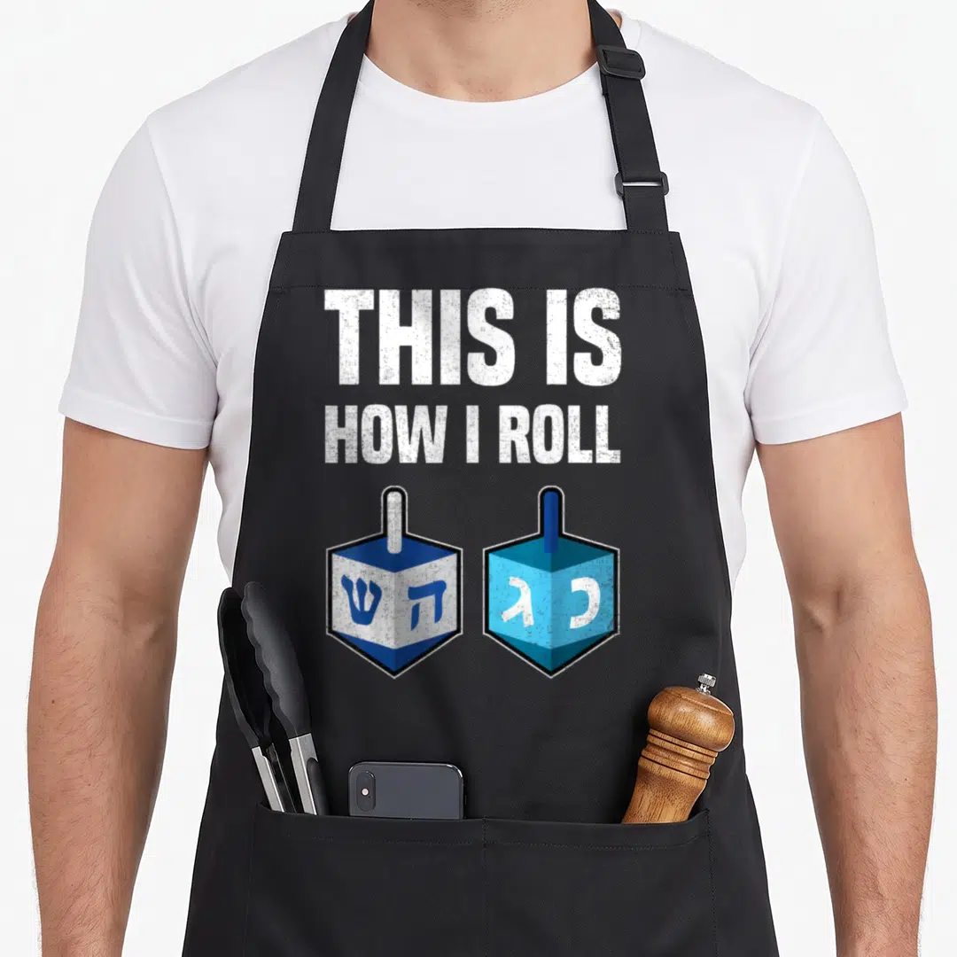 Roll hanukkah apron - THIS IS HOW I ROLL text above two blue and white dreidels with Hebrew letters — shown on a black cotton canvas apron against a white background
