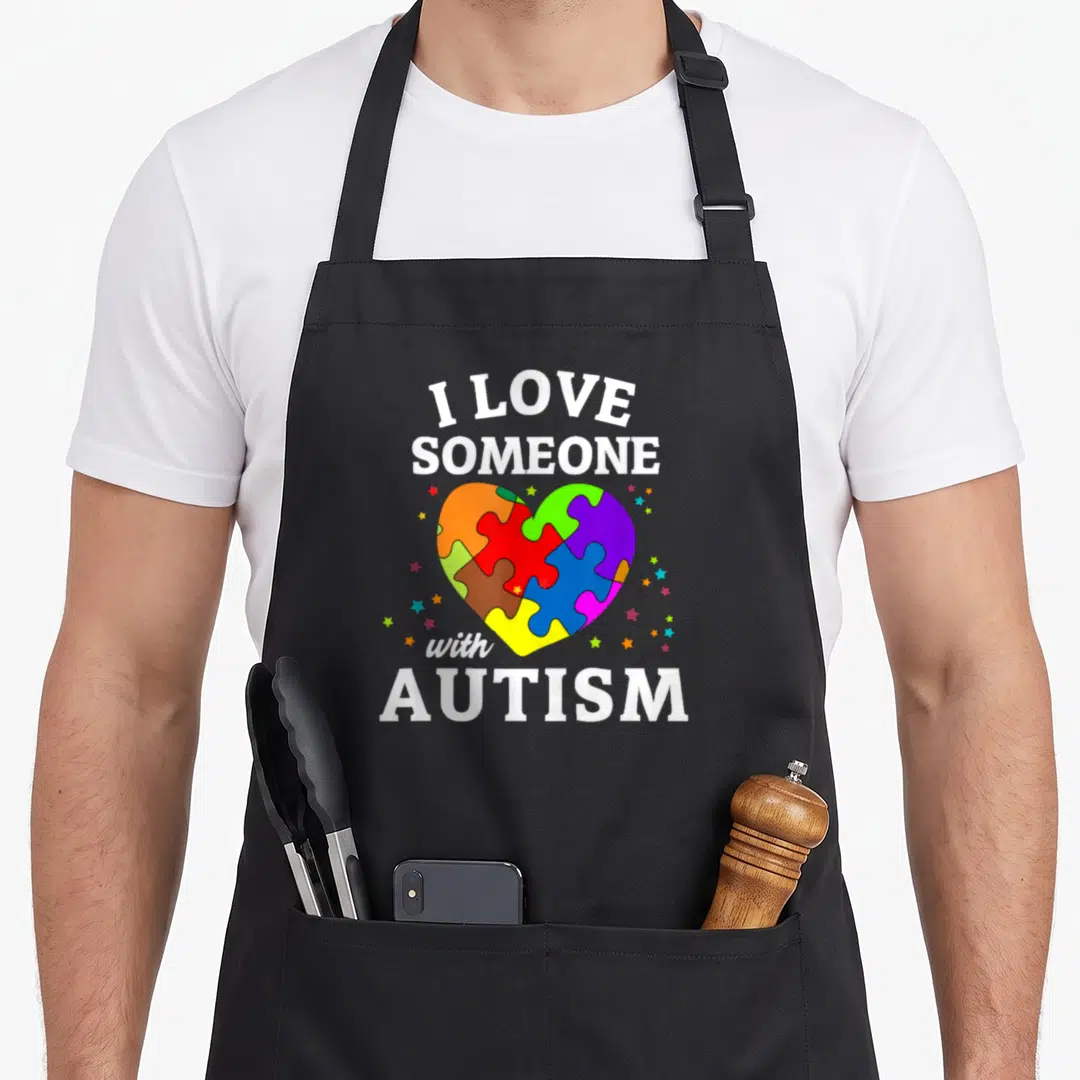 Someone autism awareness apron - I LOVE SOMEONE with AUTISM text above and below a multicolored puzzle heart surrounded — shown on a black cotton canvas apron against a white background