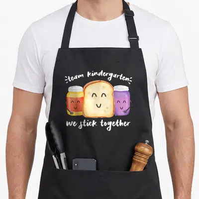 Stick kindergarten teacher apron - Watercolor bread slice between yellow and purple jars with smiling faces and white — shown on a black cotton canvas apron against a white background