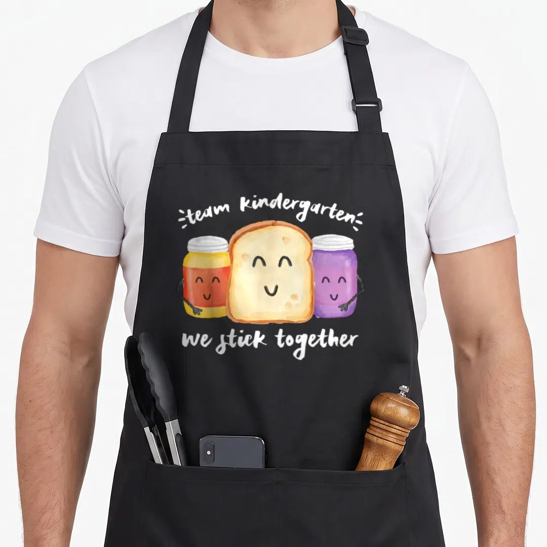Stick kindergarten teacher apron - Watercolor bread slice between yellow and purple jars with smiling faces and white — shown on a black cotton canvas apron against a white background