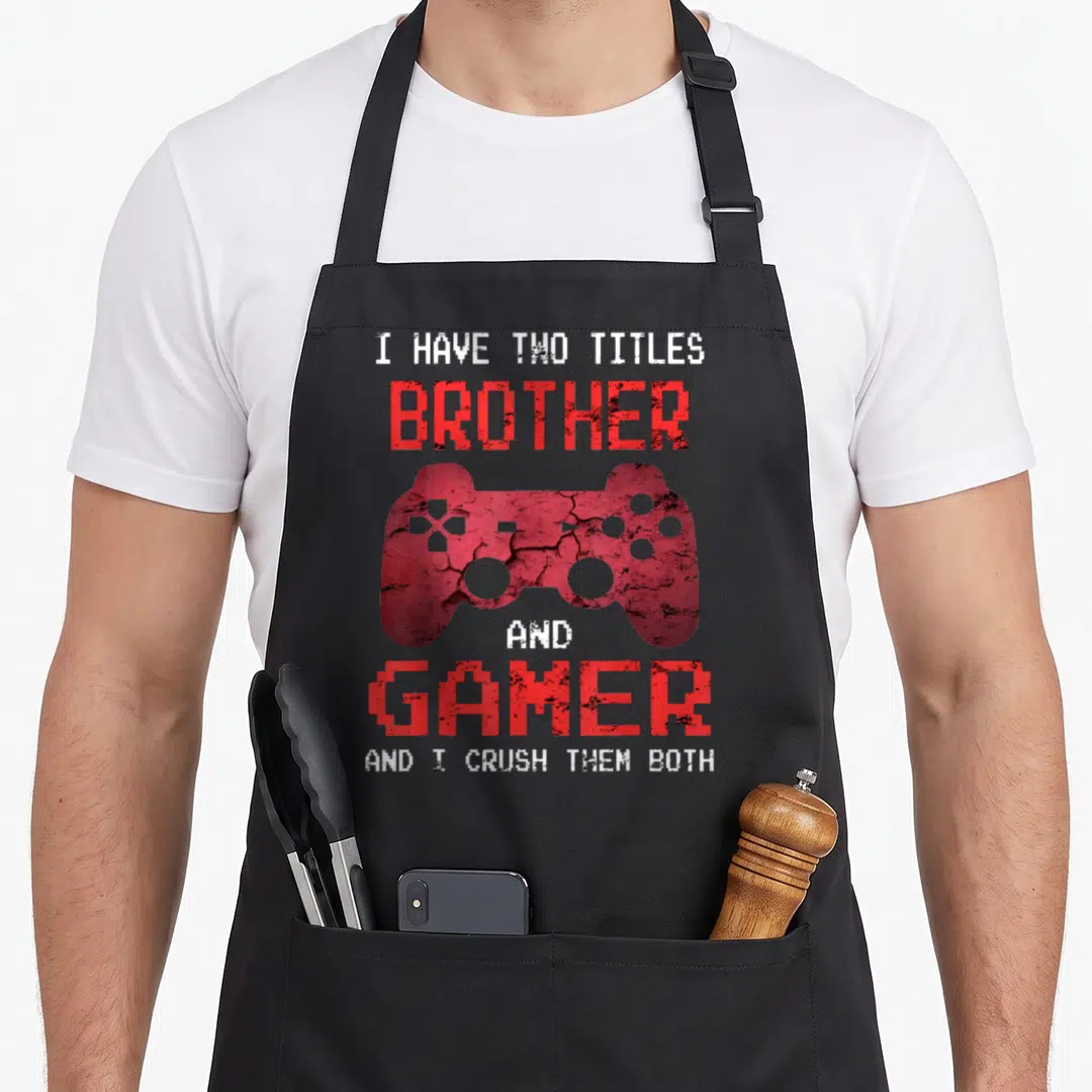 Gamer dad and son aprons - Red distressed game controller surrounded by white text reading I have two titles brother — shown on a black cotton canvas apron against a white background