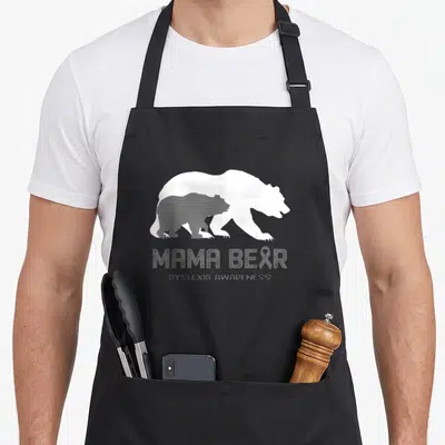 Dyslexia mama bear apron - White and grey bear silhouettes with MAMA BEAR text and ribbon in R — shown on a black cotton canvas apron against a white background