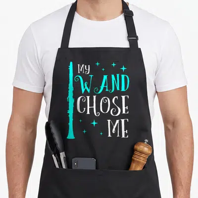 Chose clarinet apron - Cyan clarinet silhouette beside stacked white and cyan text reading my wand chose me with small — shown on a black cotton canvas apron against a white background