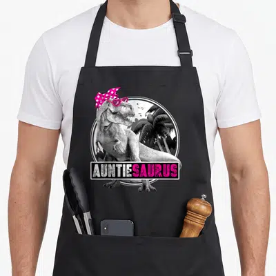 Auntiesaurus dinosaur apron - T-Rex wearing pink bow and sunglasses with AUNTIESAURUS text banner — shown on a black cotton canvas apron against a white background