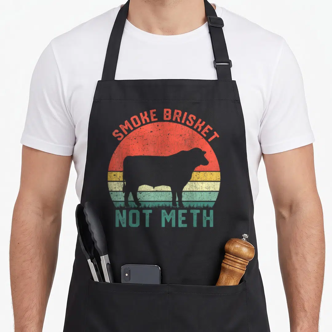 Smoke meth brisket apron - Grey cow silhouette over red yellow teal sunset with SMOKE BRISKET NOT METH text — shown on a black cotton canvas apron against a white background